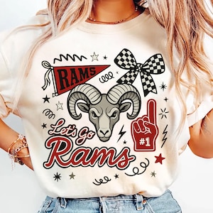 May include: Cream-colored t-shirt with a graphic design featuring a ram's head, a pennant that says "RAMS", and the text "Let's Go Rams". The design includes a foam finger, stars, and a checkered bow. The shirt is a fan item.