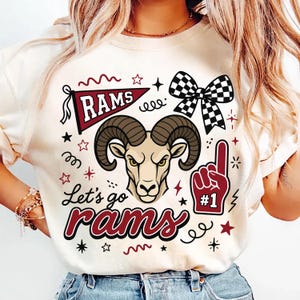 Könnte beinhalten: Ein cremefarbenes T-Shirt mit einer Grafik eines Widderkopfes, einem kastanienbraunen Wimpel mit dem Text RAMS, einer schwarz-weiß karierten Schleife und einem kastanienbraunen Schaumstofffinger mit dem Text #1. Der Text Let's go rams ist in Schreibschrift gedruckt.