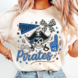 May include: Cream-colored t-shirt with a pirate-themed graphic. The design includes a skull wearing a pirate hat, crossed swords, and the words "Let's go Pirates" in blue. Additional elements include a foam finger and stars.