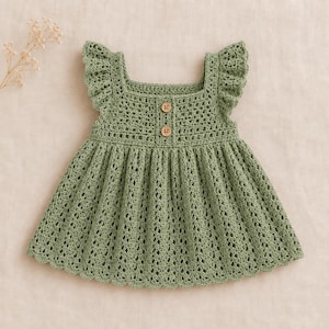 May include: A sage green sleeveless dress featuring a square neckline, ruffled shoulder straps, and a textured crochet pattern. The bodice includes two small, round, light brown wooden buttons centered vertically below the neckline.