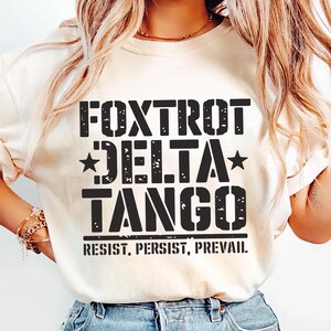 May include: A white t-shirt with black text that reads "FOXTROT DELTA TANGO RESIST, PERSIST, PREVAIL."