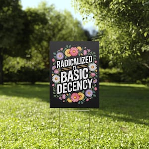 May include: A black yard sign with the text "RADICALIZED BY BASIC DECENCY" surrounded by a floral wreath of pink, yellow, and white flowers. The sign is displayed on a metal stake in a grassy garden.