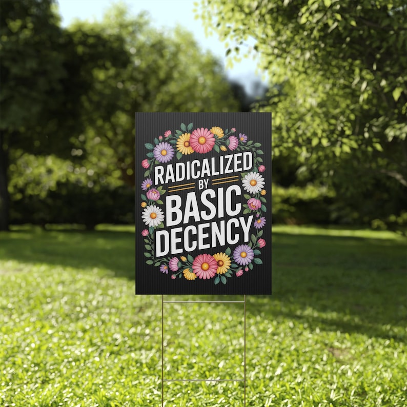 May include: A black yard sign with the text "RADICALIZED BY BASIC DECENCY" surrounded by a floral wreath of pink, yellow, and white flowers. The sign is displayed on a metal stake in a grassy garden.