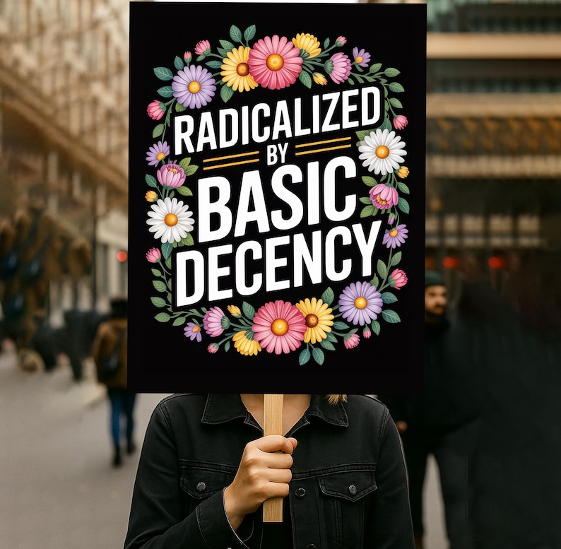 May include: A black sign with the words "RADICALIZED BY BASIC DECENCY" in white, encircled by a floral wreath of pink, yellow, purple, and white flowers. The sign is held by a person, with a blurred street scene in the background.