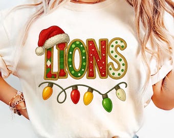 以下が含まれることがあります： クリスマスカラーで「LIONS」の文字がプリントされたクリーム色のTシャツ。「L」は赤色で、緑と金のひし形模様で、サンタ帽が被せられています。「O」と「S」は緑色で白い水玉模様です。カラフルなクリスマスライトのストリングが下にあります。