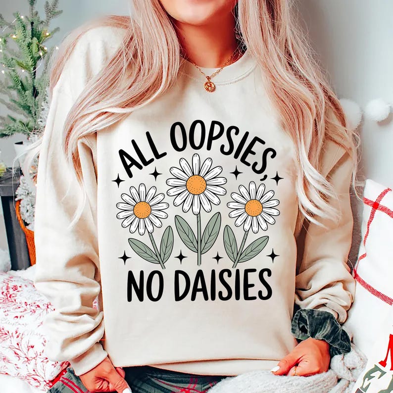 Puede incluir: Una sudadera color crema con el texto "ALL OOPSIES, NO DAISIES" en negro. El dise&ntilde;o presenta tres margaritas blancas con centros amarillos y hojas verdes. La sudadera es de estilo casual y c&oacute;modo.