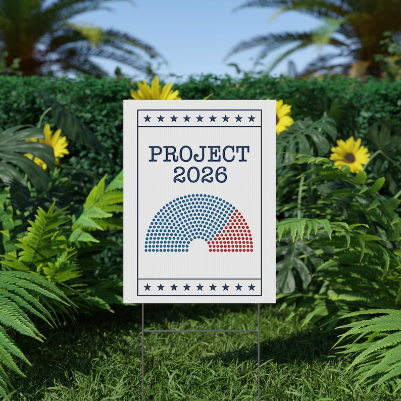 May include: A white yard sign with the text "PROJECT 2026" and a graphic of a half-circle divided into blue and red sections. The sign is supported by a metal stake and set in a grassy area with green foliage and yellow flowers.