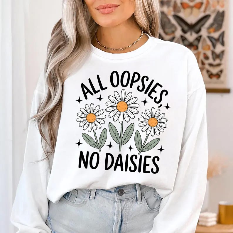 Puede incluir: Sudadera blanca con el texto negro "ALL OOPSIES NO DAISIES". El dise&ntilde;o presenta tres margaritas con p&eacute;talos blancos, centros amarillos y hojas verdes. Es una prenda superior informal de manga larga.