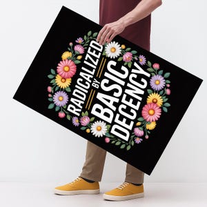 May include: A black rectangular sign featuring the text "RADICALIZED BY BASIC DECENCY" in white, encircled by a floral wreath of pink, yellow, purple, and white flowers. The sign is being held.