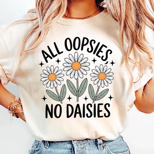 Puede incluir: Camiseta color crema con el texto negro "ALL OOPSIES NO DAISIES" y tres ilustraciones de margaritas. Las margaritas tienen p&eacute;talos blancos, centros naranjas y tallos y hojas verdes. Peque&ntilde;os acentos de estrellas negras rodean el dise&ntilde;o floral.