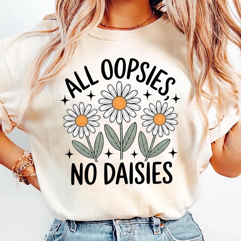 Puede incluir: Camiseta color crema con el texto negro "ALL OOPSIES NO DAISIES" y tres ilustraciones de margaritas. Las margaritas tienen p&eacute;talos blancos, centros naranjas y tallos y hojas verdes. Peque&ntilde;os acentos de estrellas negras rodean el dise&ntilde;o floral.