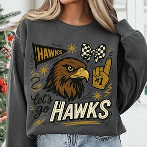 May include: Dark gray sweatshirt with a graphic design featuring a brown eagle head, a checkered bow, and the words "HAWKS" and "Let's go HAWKS". The design includes a foam finger with the number one.