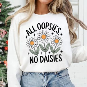 Puede incluir: Sudadera blanca de manga larga con el texto "ALL OOPSIES, NO DAISIES" en negro. El dise&ntilde;o presenta tres margaritas con p&eacute;talos blancos, centros amarillos y hojas verdes. La sudadera se combina con unos vaqueros azules.