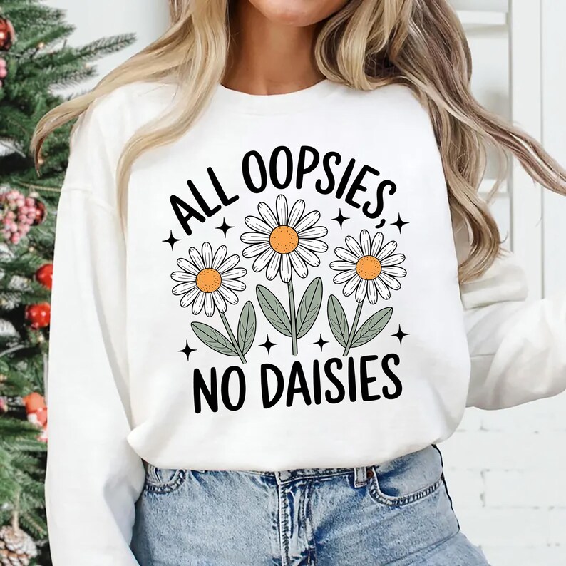 Puede incluir: Sudadera blanca de manga larga con el texto "ALL OOPSIES, NO DAISIES" en negro. El dise&ntilde;o presenta tres margaritas con p&eacute;talos blancos, centros amarillos y hojas verdes. La sudadera se combina con unos vaqueros azules.