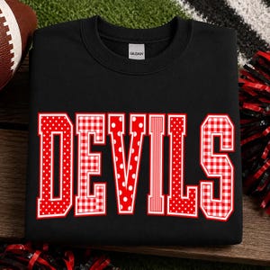 May include: A black crewneck sweatshirt featuring the word DEVILS in large, block-style letters. Each letter has a unique red and white pattern, including polka dots, gingham checks, and vertical stripes, with a white outline around each letter.