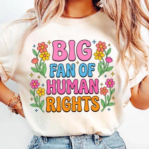 May include: Cream-colored t-shirt with the text "BIG FAN OF HUMAN RIGHTS" in a retro font. The text is surrounded by colorful flowers and leaves. The shirt is a crew neck.