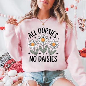 Puede incluir: Sudadera rosa claro con el texto "ALL OOPSIES, NO DAISIES" en negro. El dise&ntilde;o incluye tres margaritas con p&eacute;talos blancos y centros naranjas. Un estilo casual.