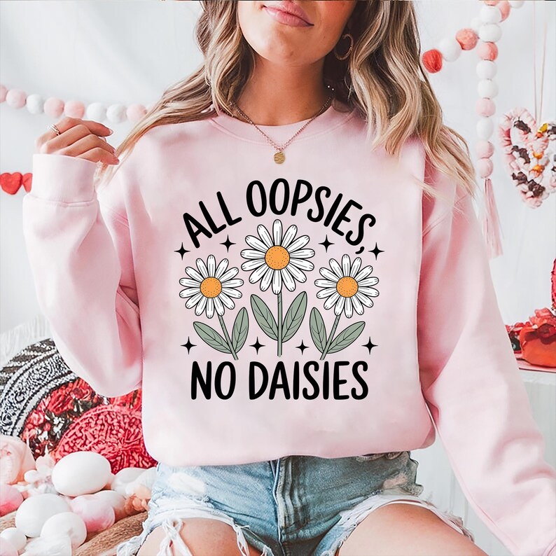 Puede incluir: Sudadera rosa claro con el texto "ALL OOPSIES, NO DAISIES" en negro. El dise&ntilde;o incluye tres margaritas con p&eacute;talos blancos y centros naranjas. Un estilo casual.