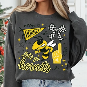 Puede incluir: Sudadera gris oscuro con un gráfico amarillo y negro. El gráfico incluye una abeja de dibujos animados, las palabras "HORNETS" y "let's go hornets", un lazo a cuadros y un dedo de espuma.