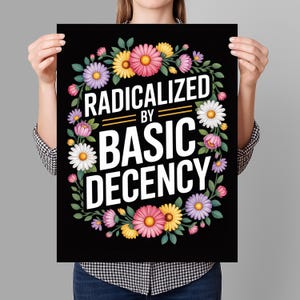 May include: Black poster with the text "RADICALIZED BY BASIC DECENCY" in white, surrounded by a floral wreath of pink, yellow, purple, and white flowers. The poster is held by a person.