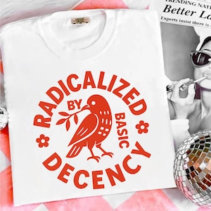 Op de afbeelding: Wit T-shirt met ronde hals en rode afbeelding. De afbeelding toont een vogel met een tak, omringd door de woorden "RADICALIZED BY BASIC DECENCY" in een cirkel. Het T-shirt is gemaakt van zacht materiaal.
