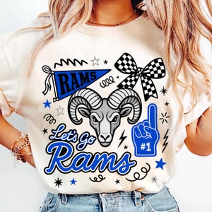 May include: A cream-colored t-shirt featuring a graphic print of a ram head, a blue pennant with the text RAMS, a blue foam finger with the text #1, a checkered bow, and the text Let's Go Rams in blue, surrounded by various star and squiggle icons.