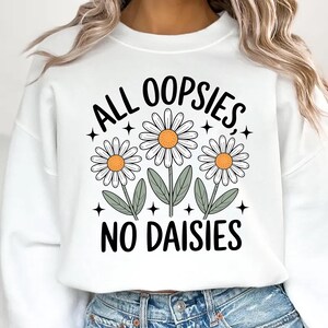 Puede incluir: Sudadera blanca con el texto negro "ALL OOPSIES, NO DAISIES". El dise&ntilde;o presenta tres margaritas con p&eacute;talos blancos, centros naranjas y hojas verdes. La sudadera es de estilo casual y c&oacute;modo.