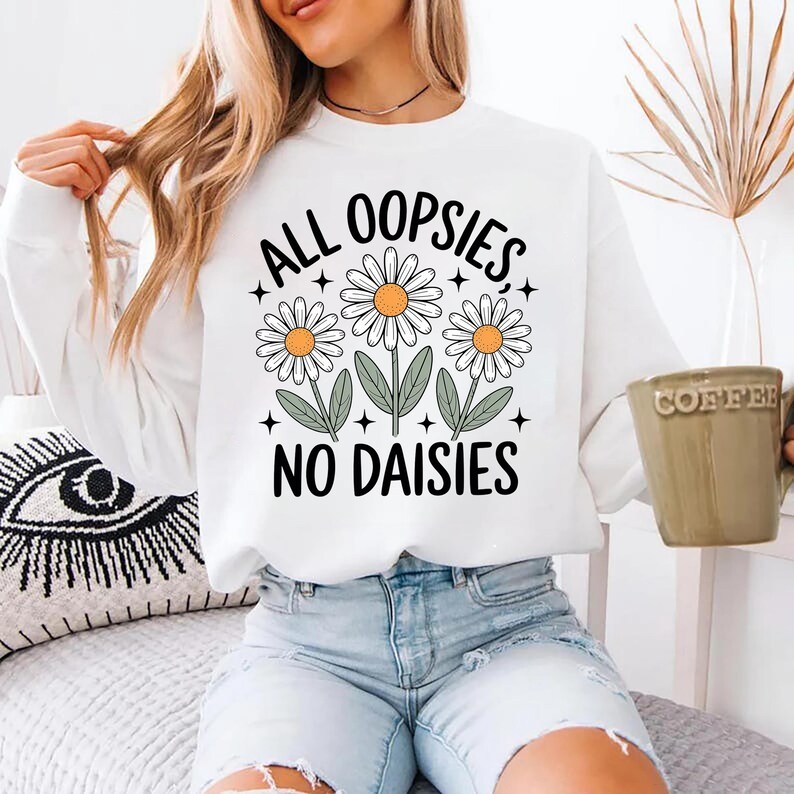 Puede incluir: Sudadera blanca con el texto "ALL OOPSIES, NO DAISIES" en negro, con tres ilustraciones de margaritas. Las margaritas tienen p&eacute;talos blancos y centros amarillos. La sudadera es de cuello redondo y manga larga.