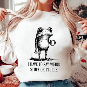 May include: White sweatshirt with a black and white illustration of a frog holding a mug. The text "I HAVE TO SAY WEIRD STUFF OR I'LL DIE" is printed below the frog. The sweatshirt is a casual style.
