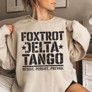 May include: A beige sweatshirt with a black graphic print that reads "FOXTROT DELTA TANGO RESIST, PERSIST, PREVAIL."