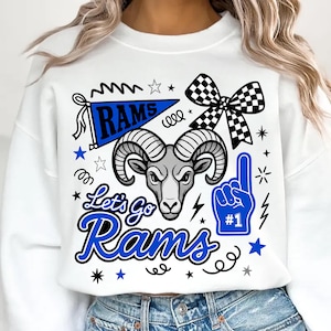Könnte beinhalten: Ein weißes Sweatshirt mit Rundhalsausschnitt und einem Grafikdruck, der einen Widderkopf, einen blauen Wimpel mit der Aufschrift "RAMS", eine schwarz-weiß karierte Schleife und einen blauen Schaumstofffinger mit "#1" zeigt. Darunter steht "Let's Go Rams".