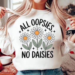 Puede incluir: Sudadera blanca con el texto negro "ALL OOPSIES NO DAISIES". El dise&ntilde;o presenta tres margaritas con p&eacute;talos blancos y centros naranjas, junto con hojas verdes y acentos de estrellas negras. Una parte superior informal con tem&aacute;tica floral.