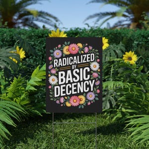 May include: A black yard sign with the text "RADICALIZED BY BASIC DECENCY" in white, surrounded by a floral wreath of pink, yellow, and white flowers. The sign is displayed on a green lawn.