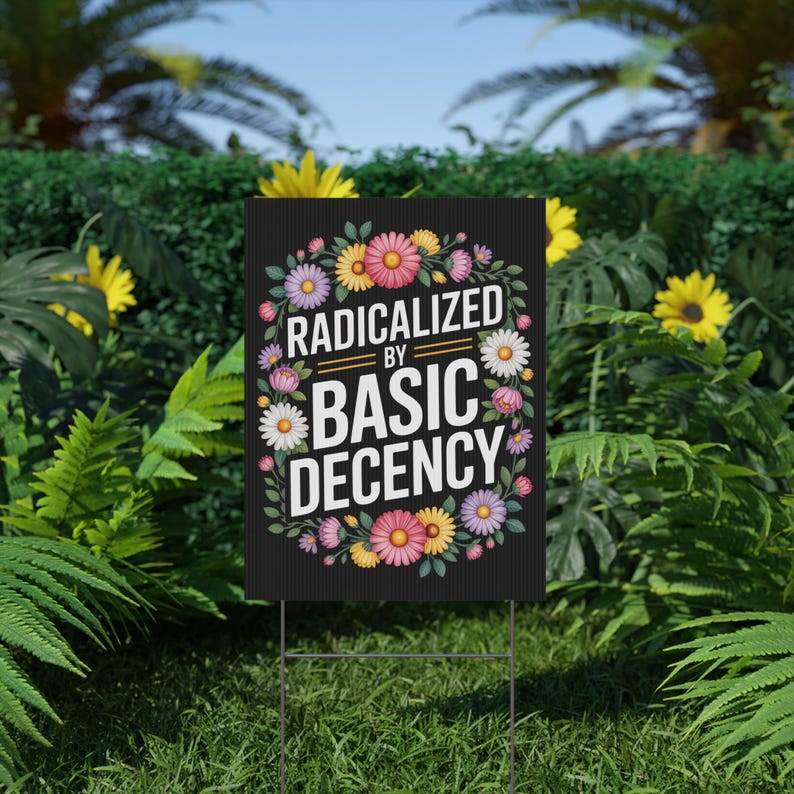 May include: A black yard sign with the text "RADICALIZED BY BASIC DECENCY" in white, surrounded by a floral wreath of pink, yellow, and white flowers. The sign is displayed on a green lawn.