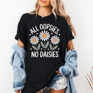 Puede incluir: Camiseta negra con el texto blanco "ALL OOPSIES, NO DAISIES". El dise&ntilde;o presenta tres margaritas con p&eacute;talos blancos y centros naranjas. Hojas verdes y peque&ntilde;as estrellas completan el gr&aacute;fico floral.