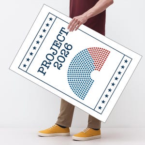 May include: A white sign with the text "PROJECT 2026" in navy blue, featuring a graphic of blue and red dots. The sign is held by a person wearing a maroon shirt, khaki pants, and yellow sneakers.