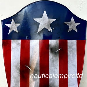 May include: A red, white, and blue shield with a distressed look. The shield has three white stars on a blue background and red and white stripes. The shield has the text "nauticalempireltd" at the bottom.
