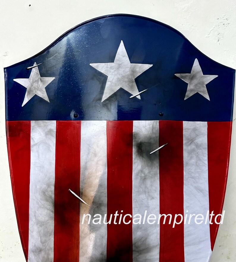 May include: A red, white, and blue shield with a distressed look. The shield has three white stars on a blue background and red and white stripes. The shield has the text "nauticalempireltd" at the bottom.
