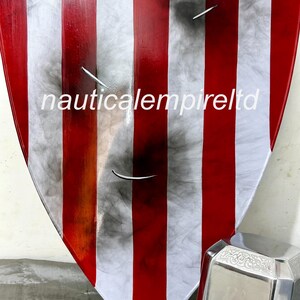 May include: A red, white, and blue shield with a distressed, weathered look. The shield is made of metal and has a raised edge. The shield is decorated with a star in the center. The shield is leaning against a silver metal object with a circular design.