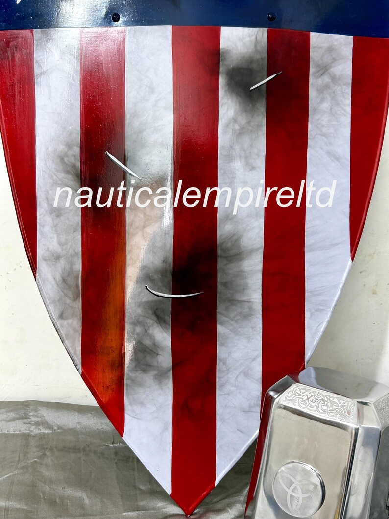 May include: A red, white, and blue shield with a distressed, weathered look. The shield is made of metal and has a raised edge. The shield is decorated with a star in the center. The shield is leaning against a silver metal object with a circular design.