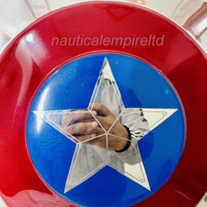 May include: A red, white and blue Captain America shield with a silver star in the centre. The shield is made of metal and has a shiny finish. The text "nauticalempireltd" is printed on the shield.