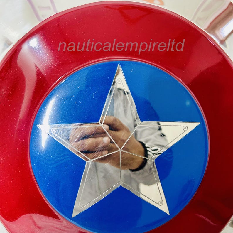 May include: A red, white and blue Captain America shield with a silver star in the centre. The shield is made of metal and has a shiny finish. The text "nauticalempireltd" is printed on the shield.