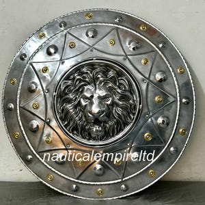 Pode incluir: Um escudo redondo prateado com uma cabeça de leão em relevo no centro. O escudo é decorado com rebites dourados ao redor da borda e em um padrão de estrela ao redor da cabeça do leão. O texto "nauticalempireltd" está impresso no escudo.