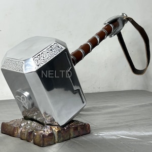 Könnte beinhalten: Ein silberfarbener Hammer mit einem braunen Ledergriff und einem Riemen. Der Hammer hat aufwendige Schnitzereien und ein Symbol auf dem Kopf. Der Hammer liegt auf einem braunen Metallständer.
