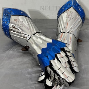 Könnte beinhalten: Paar mittelalterliche Panzerhandschuhe. Die Panzerhandschuhe bestehen aus poliertem Silbermetall mit blauen Akzenten. Das Design umfasst bewegliche Platten und dekorative Nieten. Die Panzerhandschuhe dienen zum Schutz der Hände und Handgelenke.