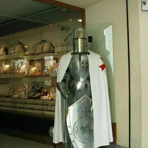May include: A full suit of silver armor with a white and red cape, a helmet with a golden cross, and a shield with a golden cross and a knight riding a horse. The armor is standing on a black base.