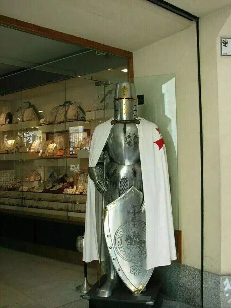 May include: A full suit of silver armor with a white and red cape, a helmet with a golden cross, and a shield with a golden cross and a knight riding a horse. The armor is standing on a black base.