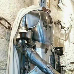May include: A silver knight's armor with a white cape, a helmet with a cross, and a shield with a cross. The armor is standing against a stone wall.