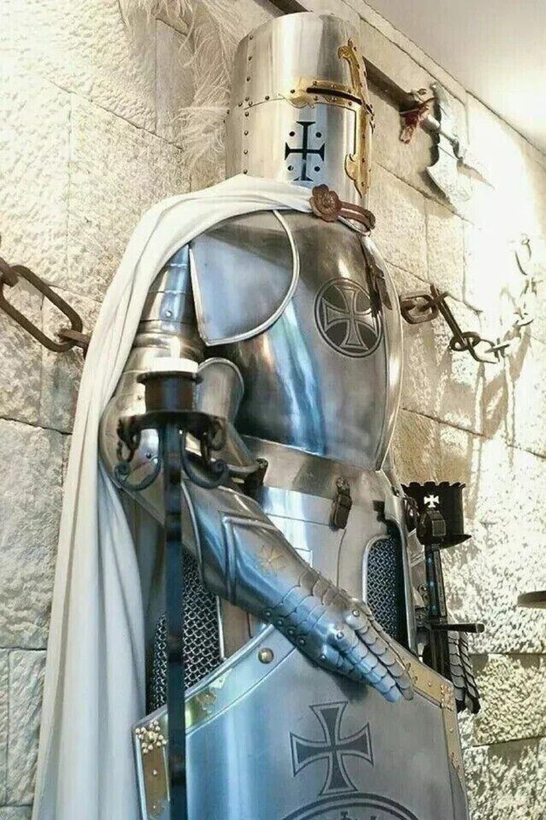 May include: A silver knight's armor with a white cape, a helmet with a cross, and a shield with a cross. The armor is standing against a stone wall.
