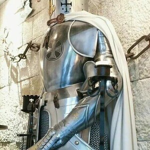 May include: A silver knight in full armor with a white cape and a black cross on the chest plate. The knight is holding a sword and a shield with a black cross.